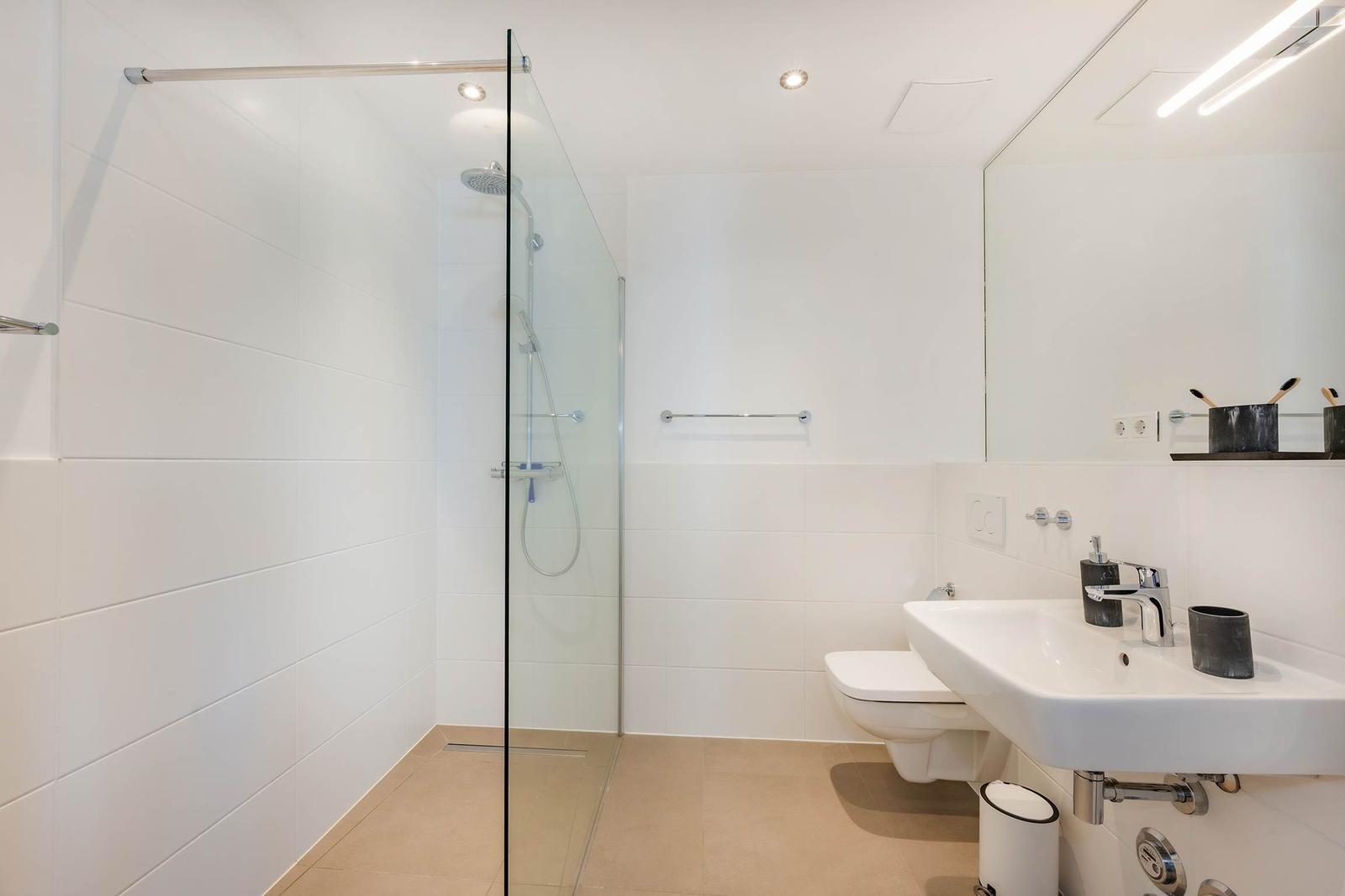Badezimmer mit Dusche, Waschbecken und Toilette. Weiße Fliesen und moderne Einrichtung.