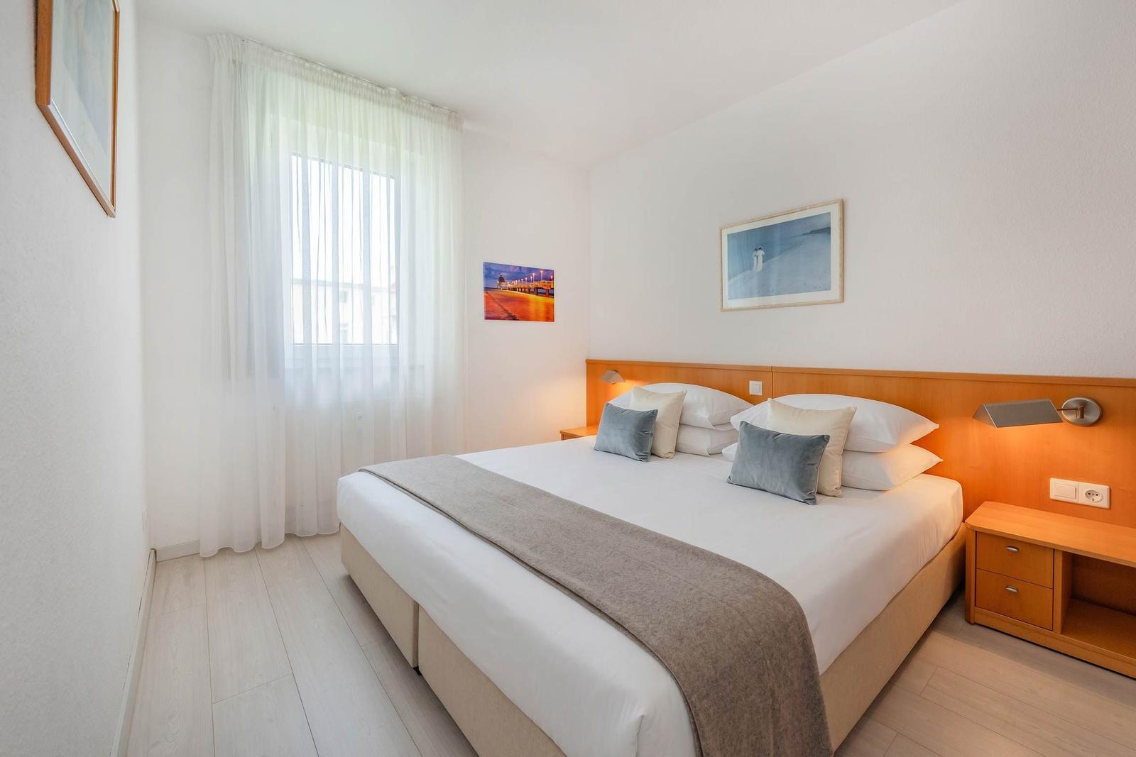 Schlafzimmer mit Doppelbett, Holzfußboden und Fenster mit weißen Vorhängen.