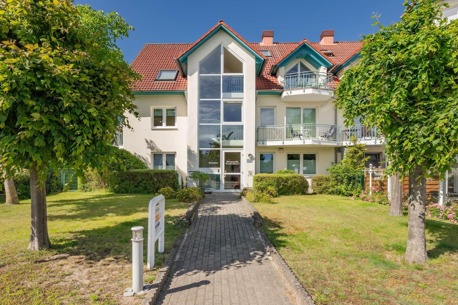 Haus mit Eingangsbereich, Balkonen und gepflegtem Garten unter blauem Himmel.