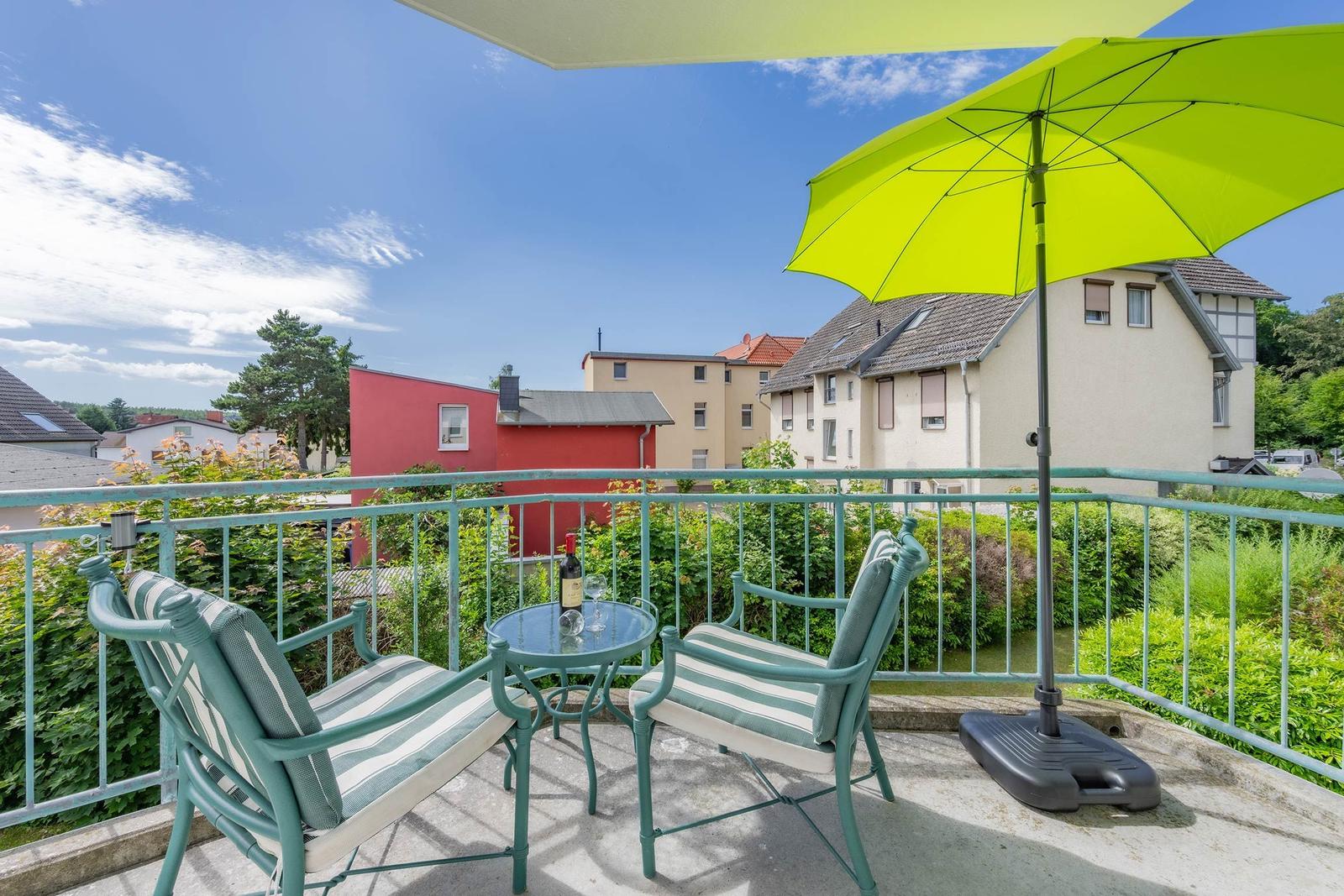 Terrasse mit Tisch, zwei Stühlen und grünem Sonnenschirm. Blick auf Wohnhäuser und Grünflächen.