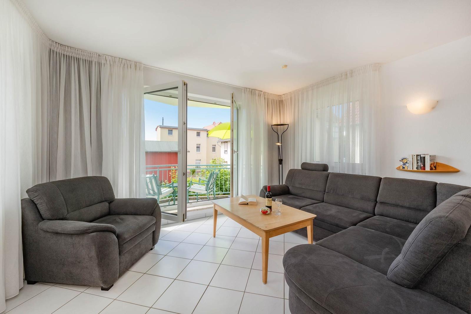 Wohnzimmer mit grauem Sofa, Tisch und Balkon. Helle Wand, weiße Fliesen.