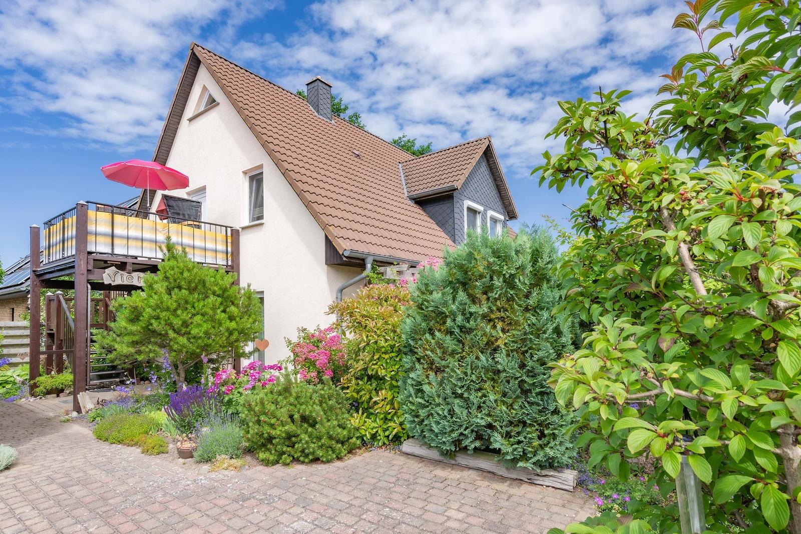 Haus mit Balkon, rotem Sonnenschirm und blühendem Garten unter blauem Himmel.
