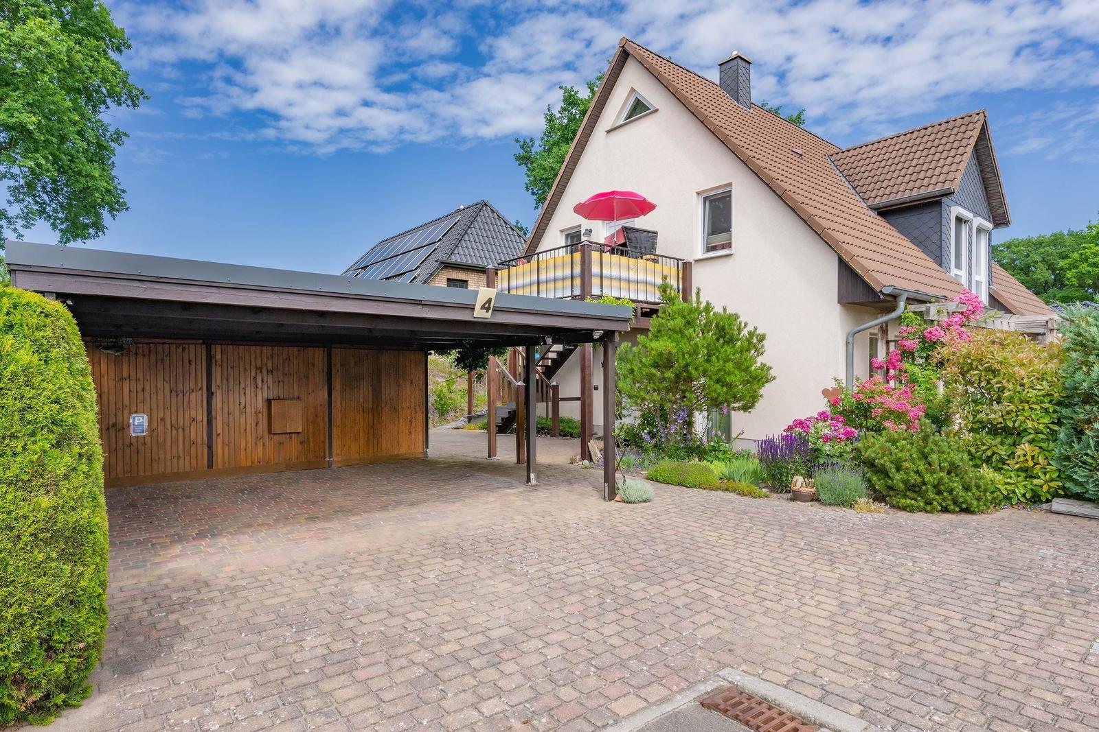 Haus mit Terrasse, rotem Sonnenschirm und gepflasterter Hof. Garten mit Blumen und Bäumen.