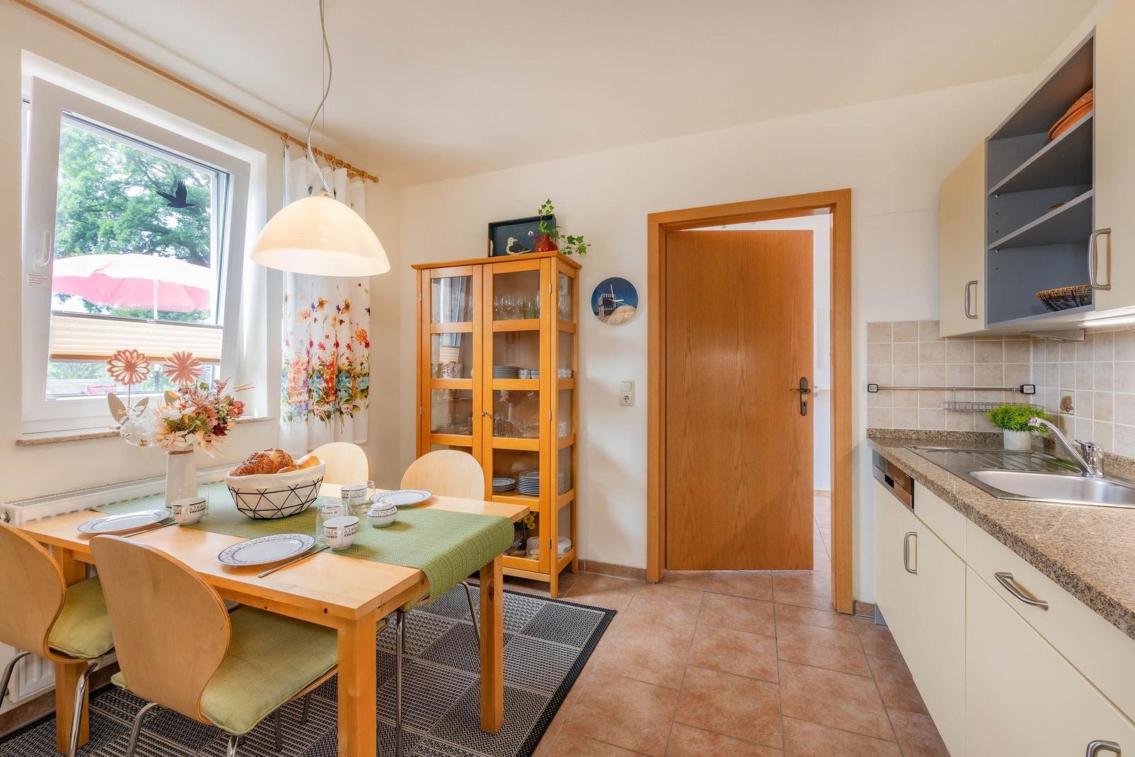 Esszimmer mit Tisch, Stühlen und Küchenbereich. Fenster mit Blick auf Garten.