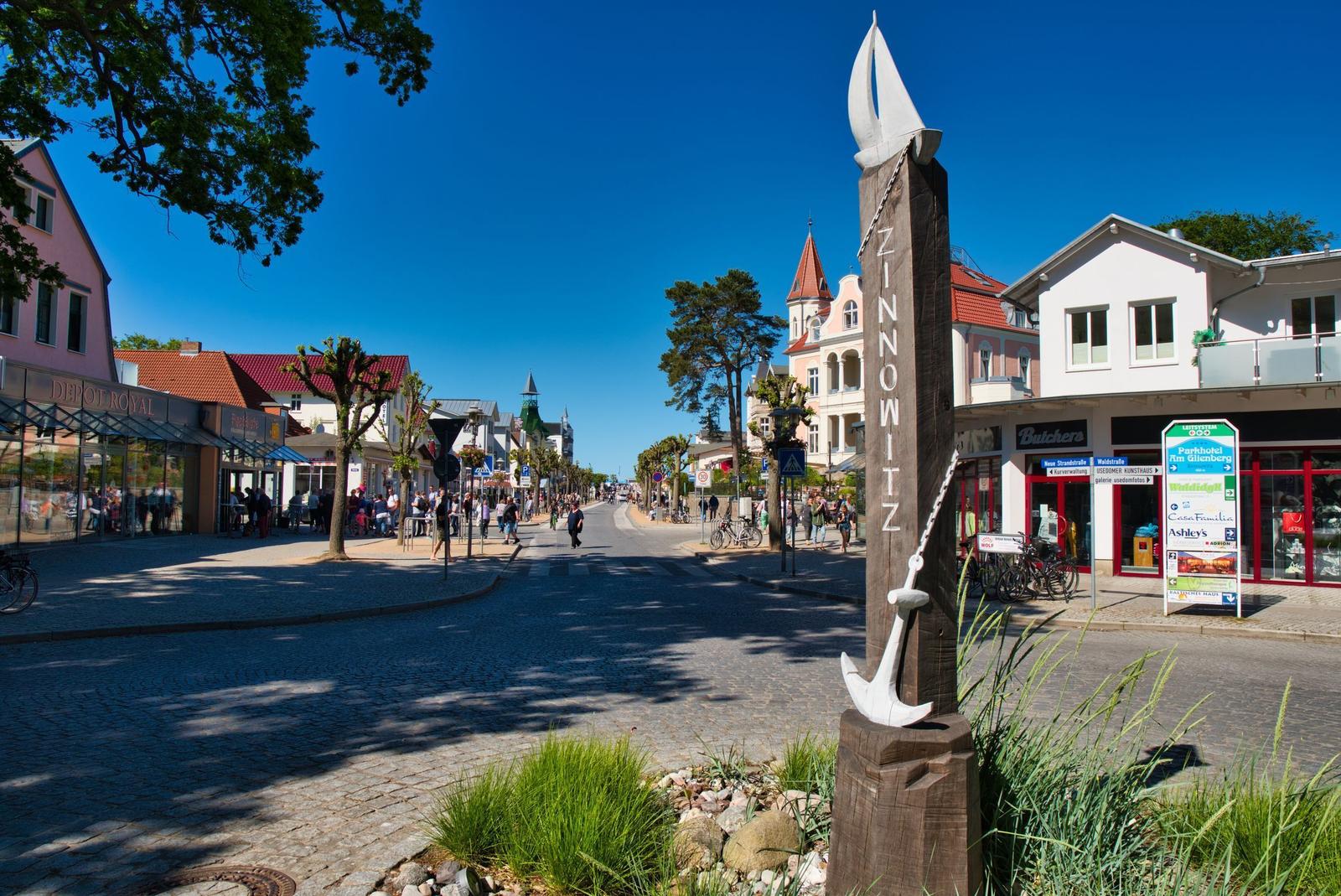 Zinnowitz Hauptstraße mit Markierungssäule, Geschäften und Fußgängern unter blauem Himmel.