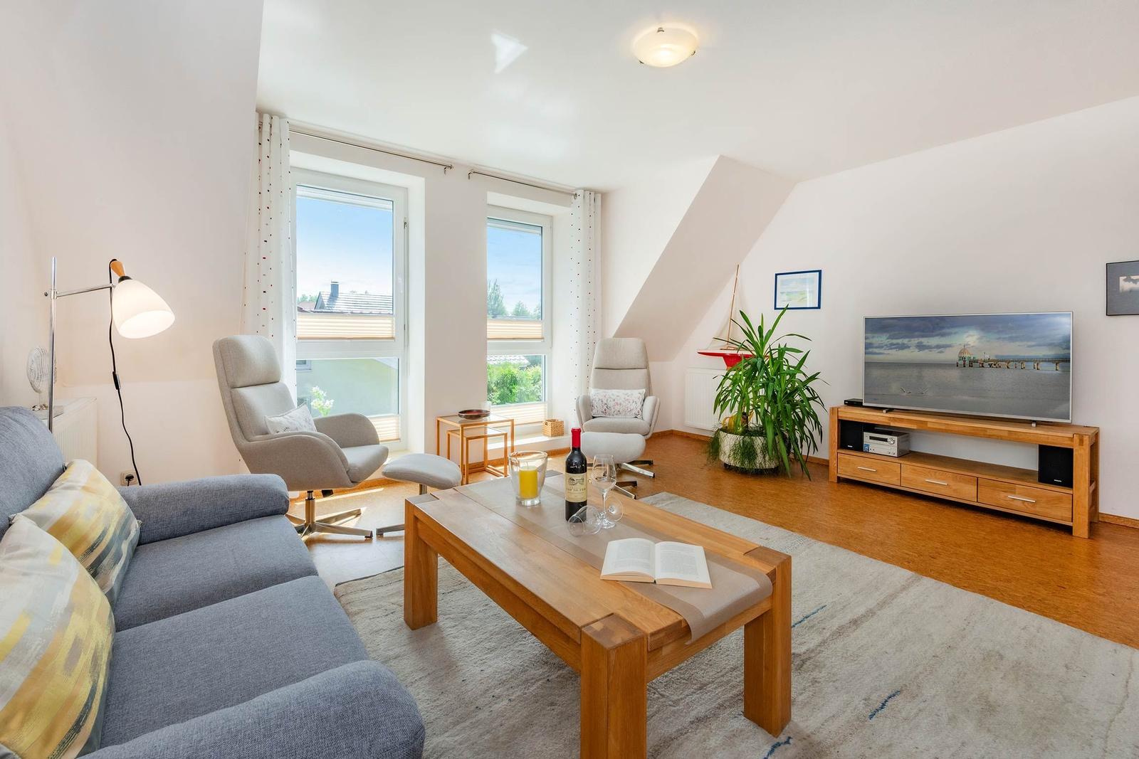 Wohnzimmer mit Sofa, zwei Sesseln, Holztisch und Fernseher. Fenster mit Blick nach draußen.