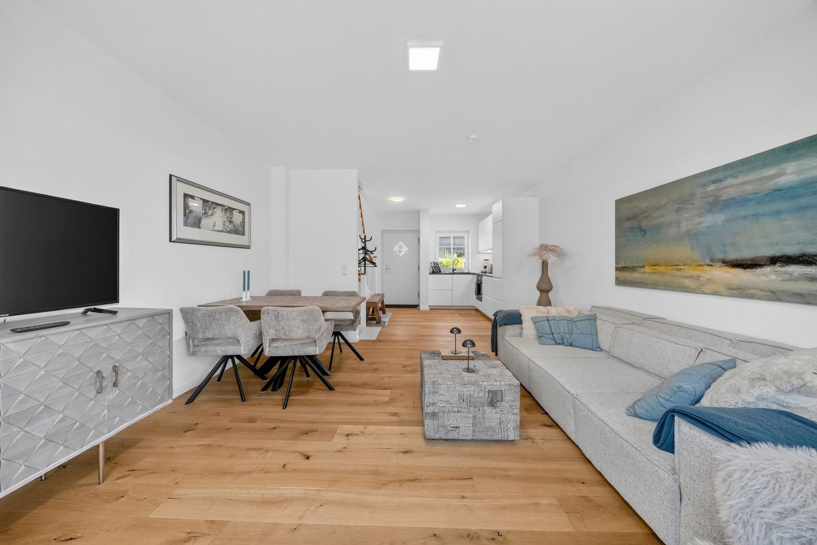 Wohn- und Essbereich mit Sofa, Esstisch und Fernseher. Holzboden und weiße Wände.
