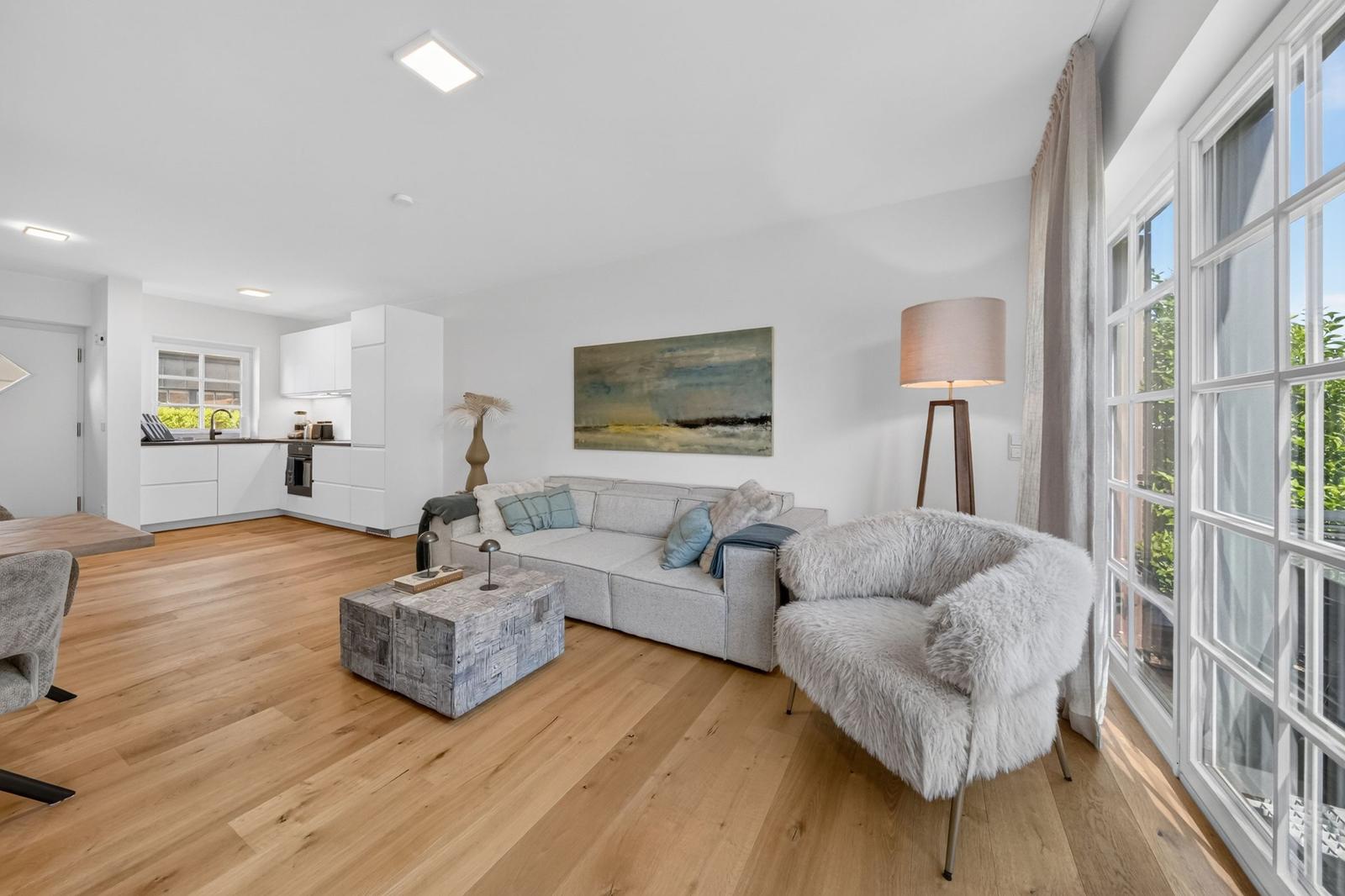 Wohnzimmer mit Sofa, Fellstuhl und Küchenbereich. Holzboden und große Fenster.