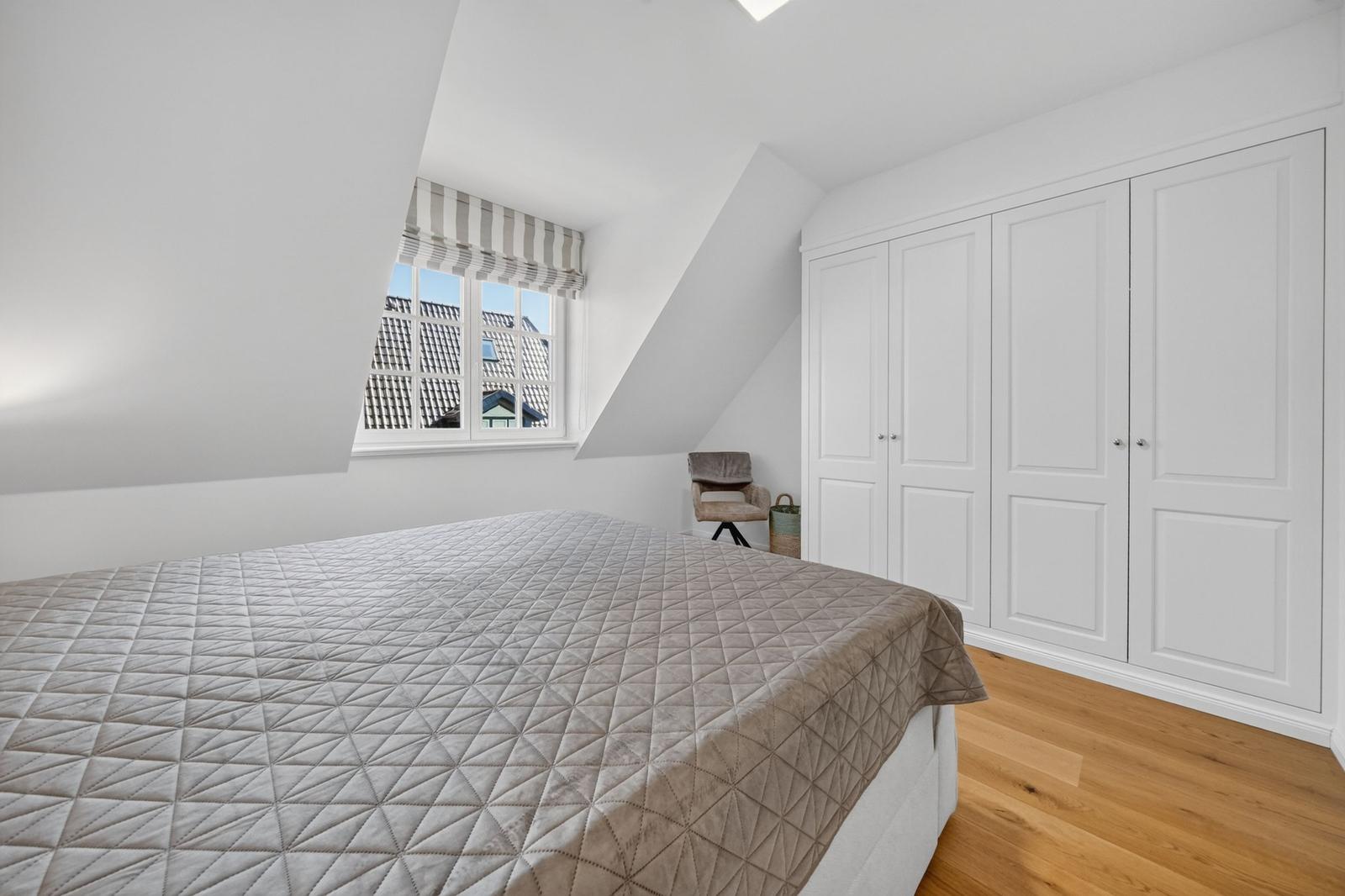 Schlafzimmer mit Dachschrägen, Bett, Kleiderschrank und Fenster mit Blick auf Dächer.
