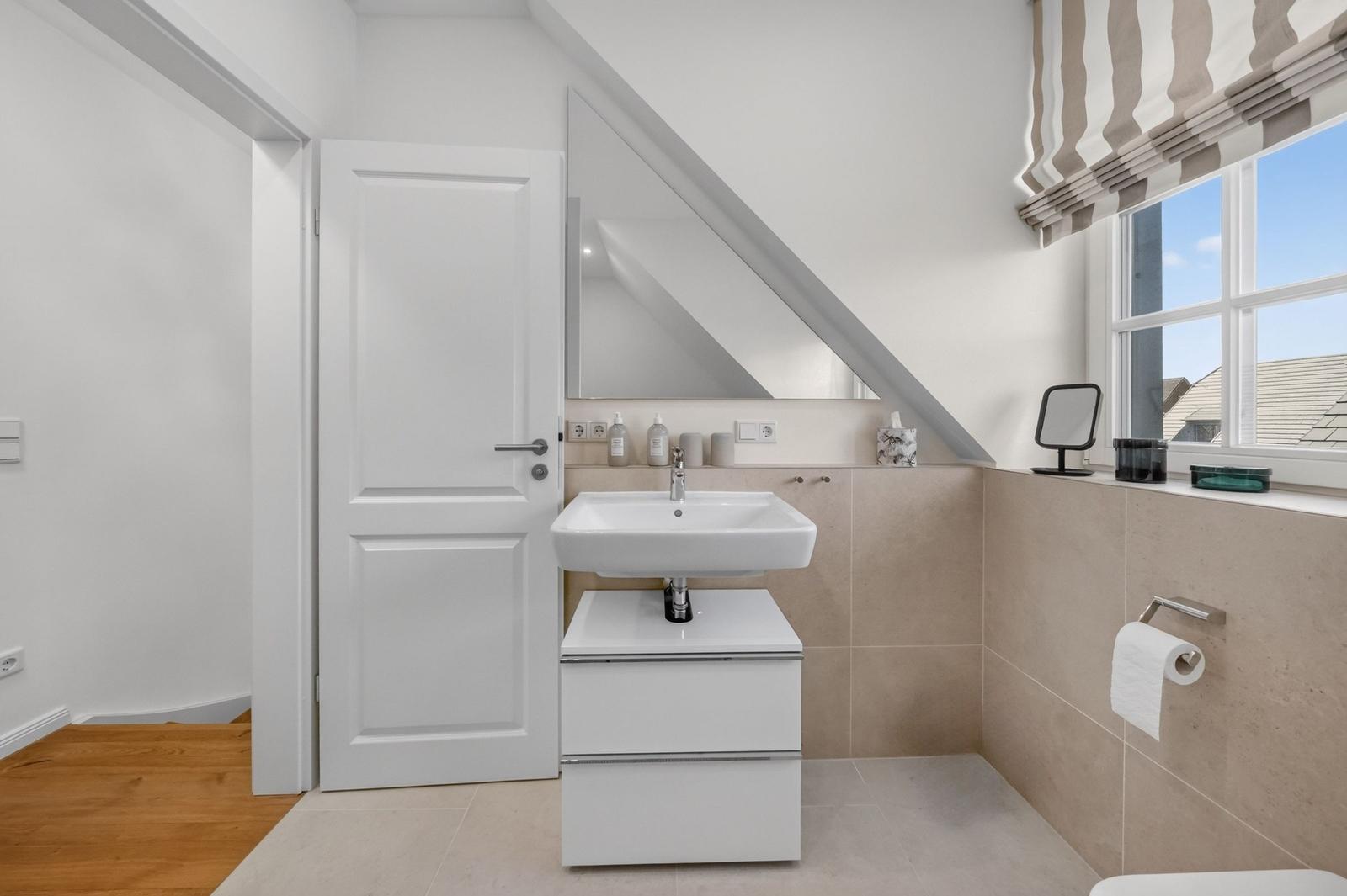 Badezimmer mit Waschbecken, Spiegel und Fenster. Holzboden und beige Fliesen.