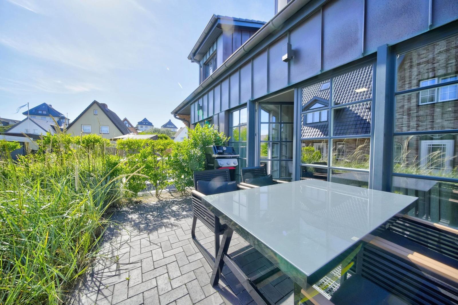 Terrasse mit Tisch, Stühlen und Grill. Blick auf grüne Pflanzen und benachbarte Häuser.