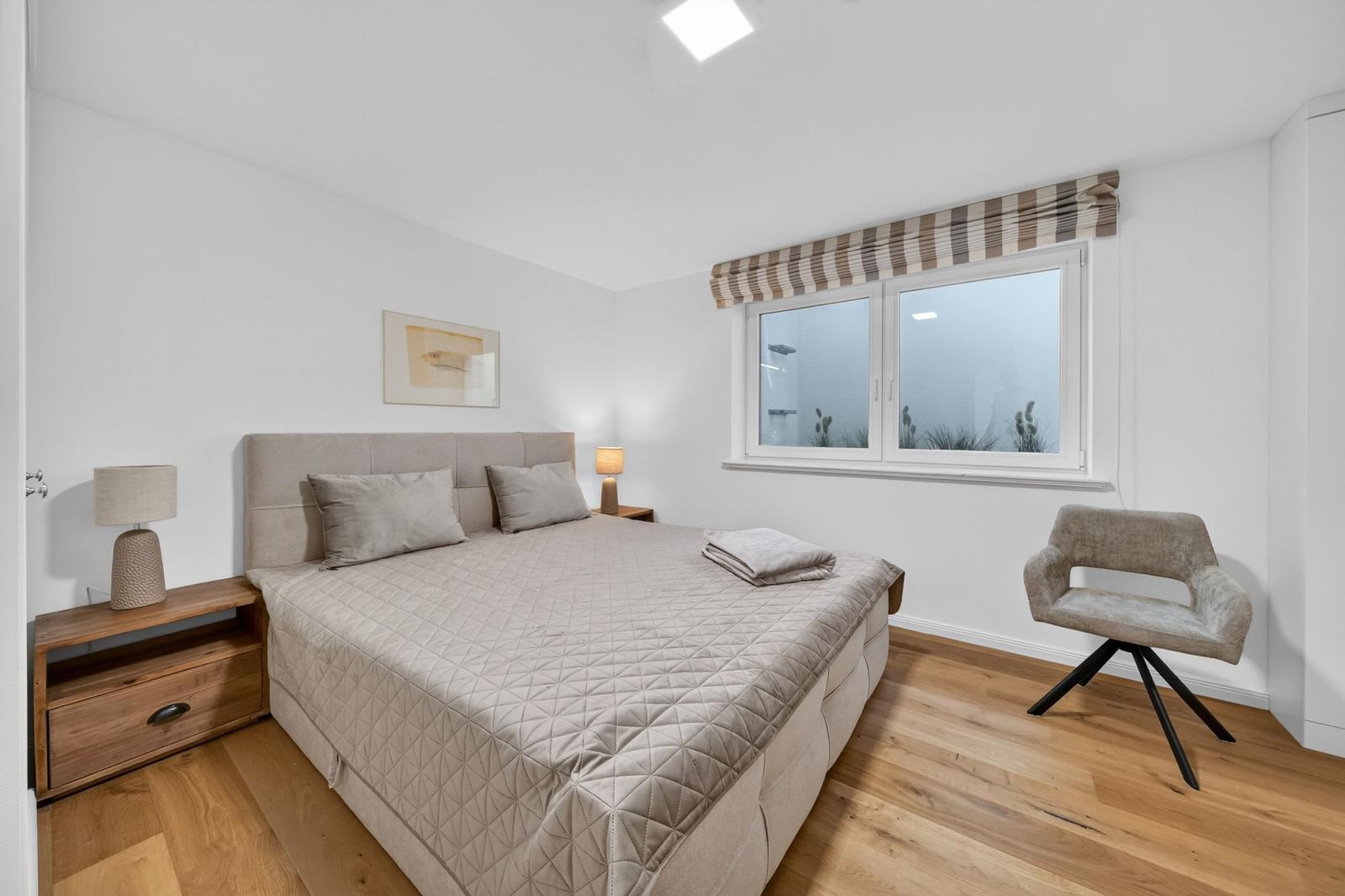 Schlafzimmer mit Bett, Nachttisch, Stuhl und Fenster.
