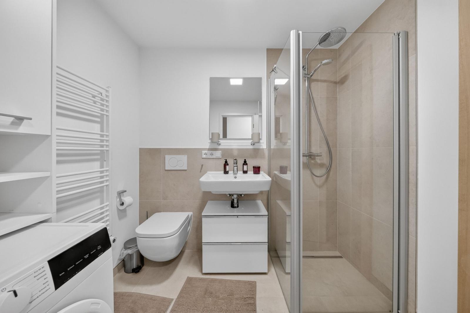 Badezimmer mit Dusche, Waschbecken und Toilette. Links steht eine Waschmaschine.