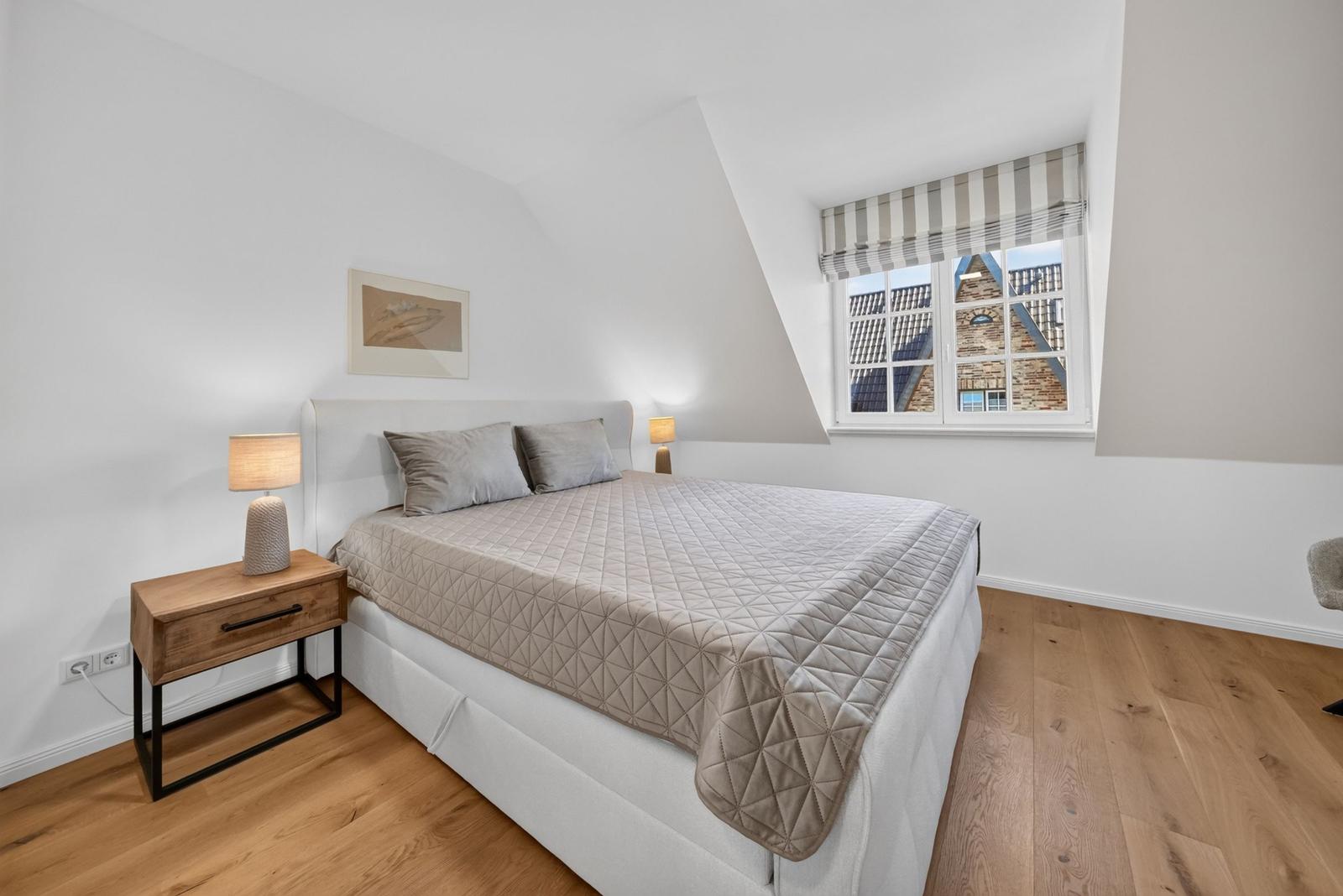 Schlafzimmer mit Dachschräge, Bett, Nachttisch und Fenster mit Blick auf Gebäude.