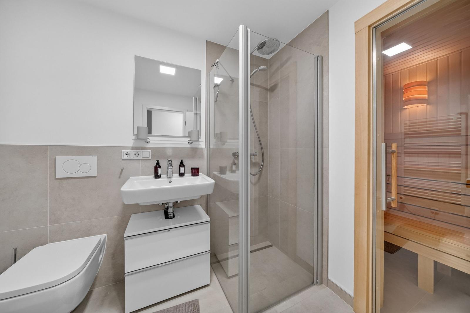 Badezimmer mit Dusche, Waschbecken und Toilette. Nebenraum ist eine Sauna.