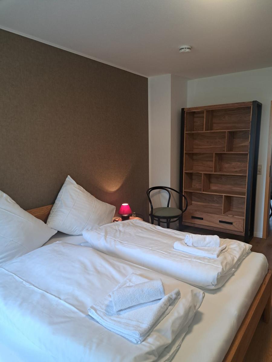Zimmer mit Doppelbett, Holzregal und Nachttisch mit Lampe.
