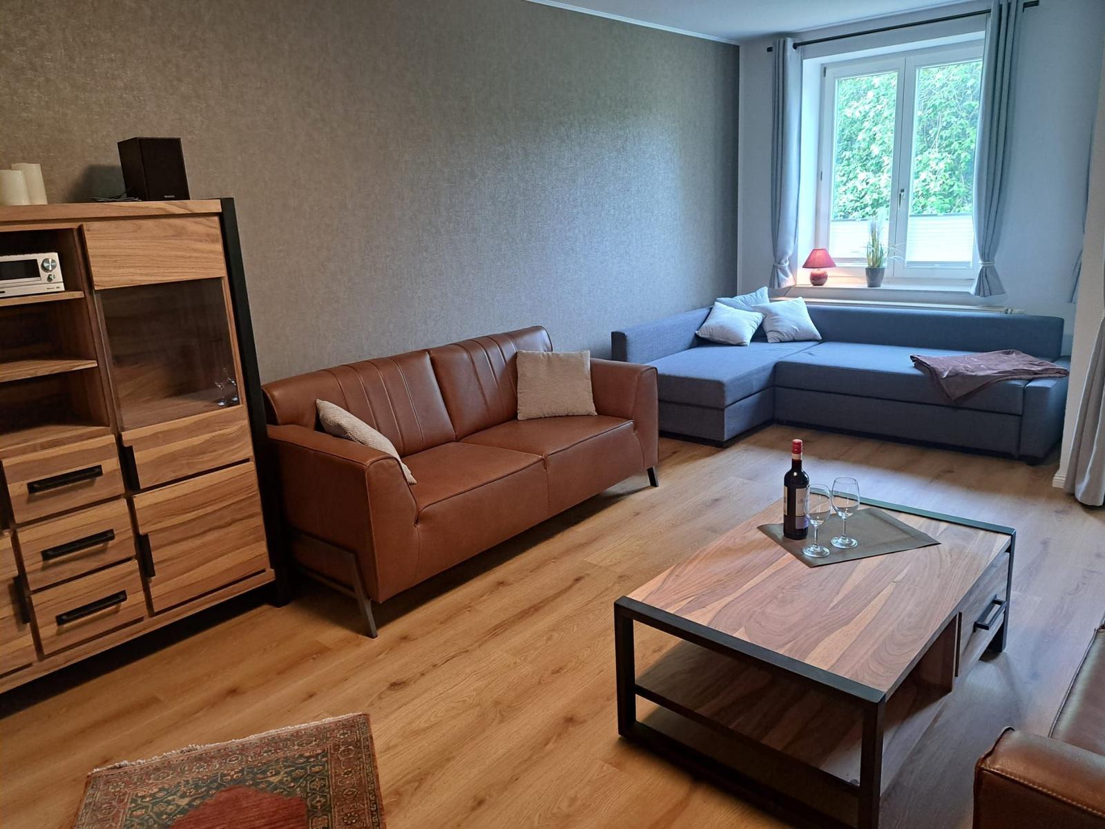 Wohnzimmer mit braunem Ledersofa, blauem Ecksofa, Holztisch und Fenster mit Blick auf Bäume.