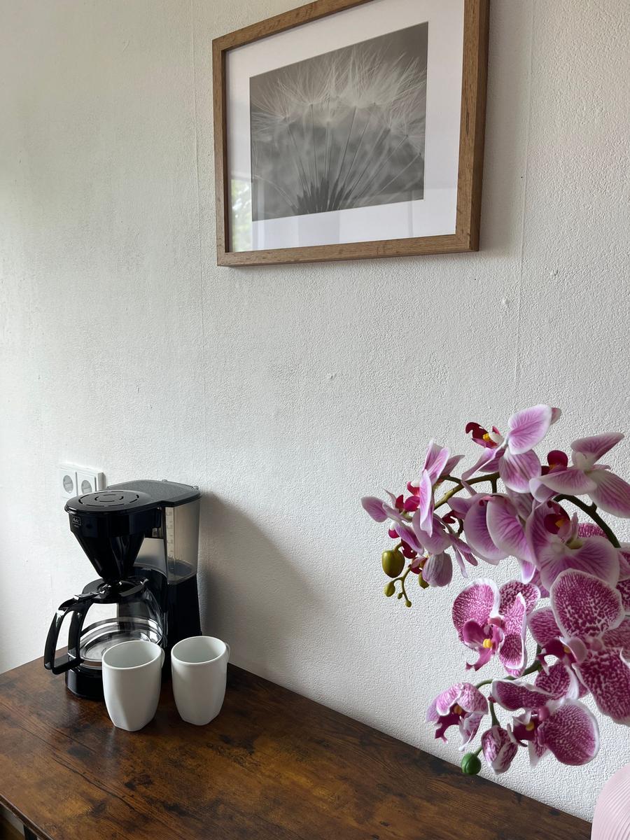 Eine Kaffeemaschine mit zwei Tassen auf einem Holztisch neben einer Orchidee und einem Bild an der Wand.