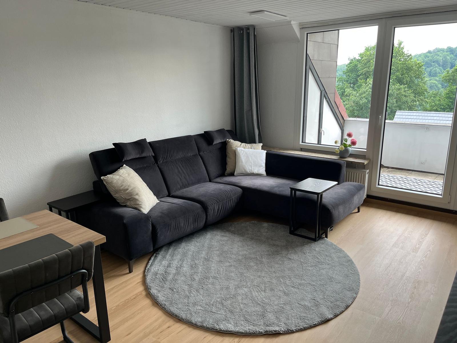 Ein Wohnzimmer mit dunklem Sofa, runden Teppich und Blick auf Balkon.