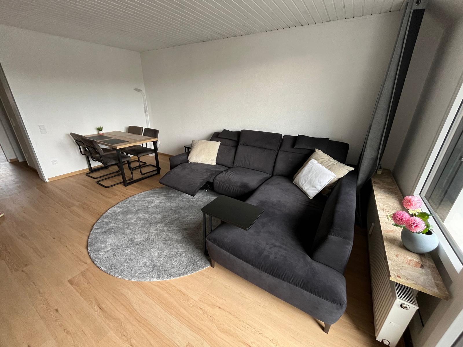 Ein Wohnzimmer mit grauem Sofa, runden Teppich und Esstisch im Hintergrund.
