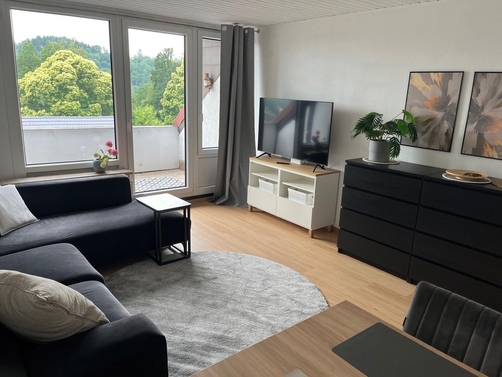 Wohnzimmer mit Sofa, Fernseher und Balkonblick auf grüne Bäume.