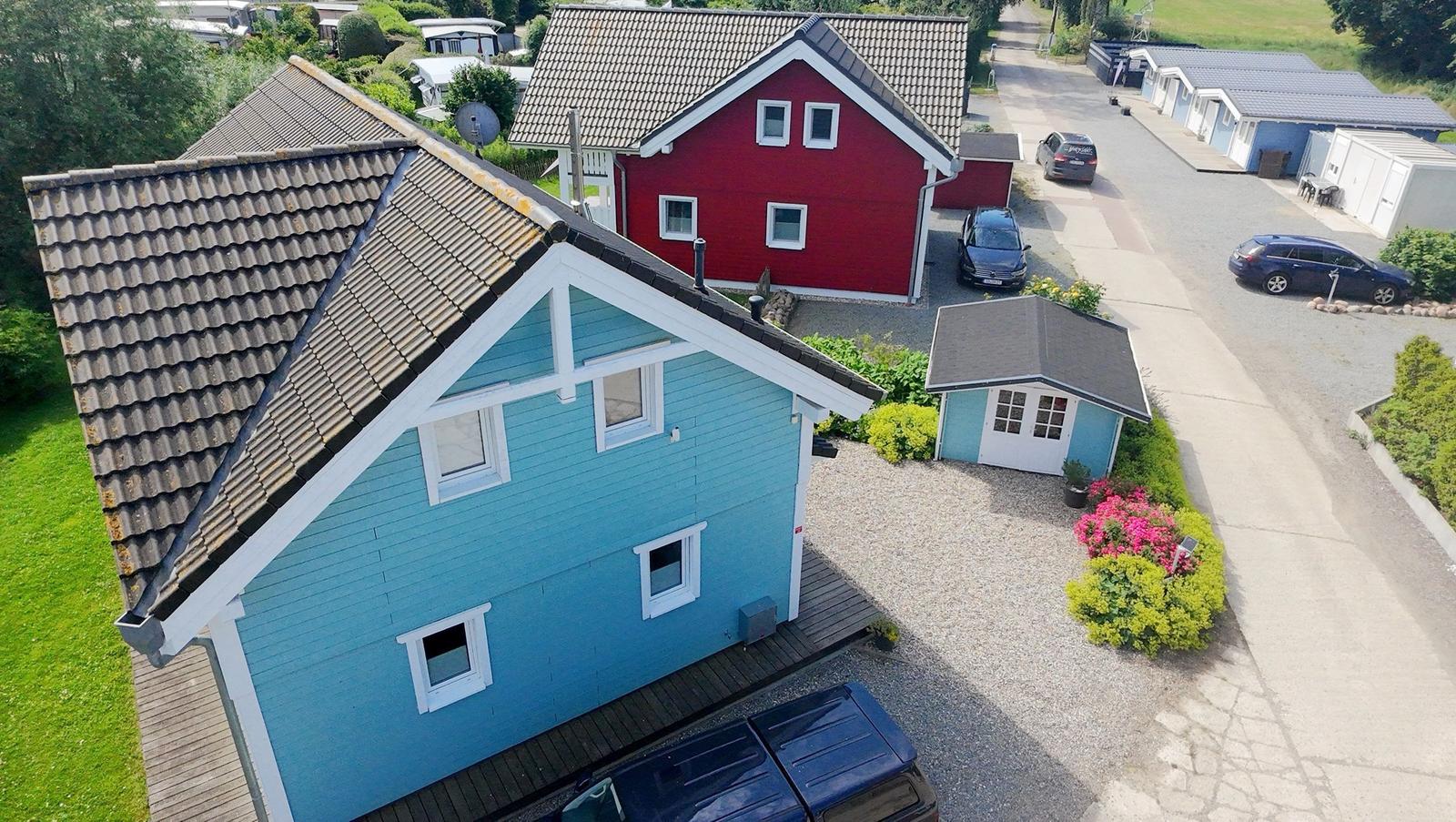 Blau gestrichenes Haus mit Dach und Garten. Rotes Haus im Hintergrund. Mehrere Autos parken.