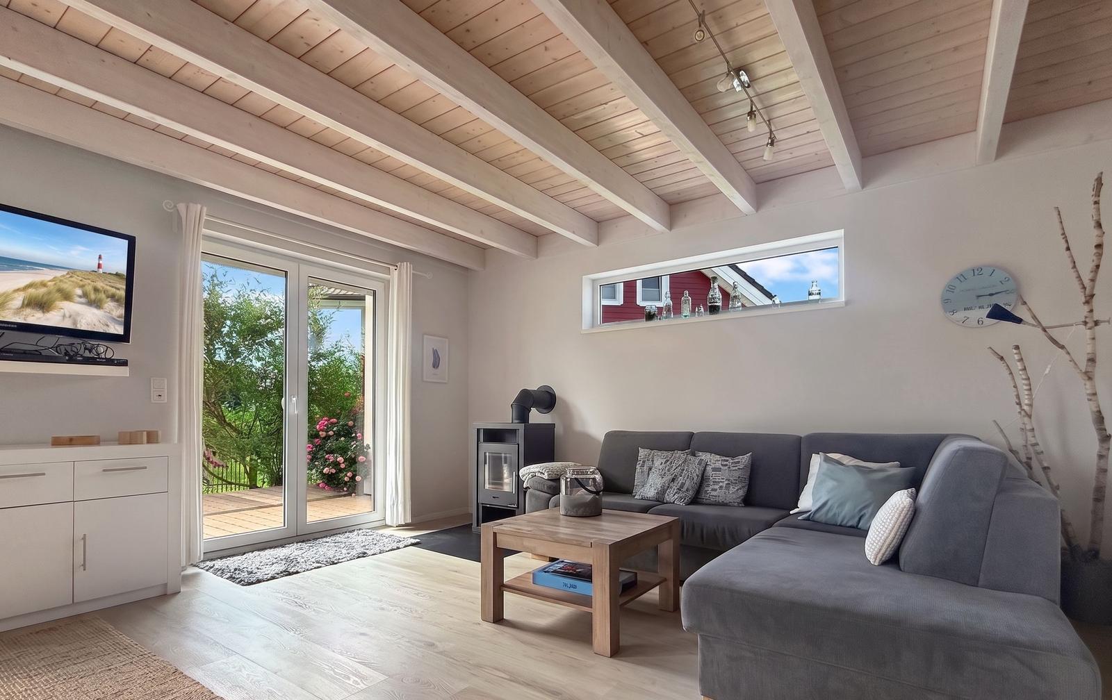 Wohnzimmer mit grauem Sofa, Holztisch und Fernseher. Durchgehende Glastür führt auf die Terrasse.