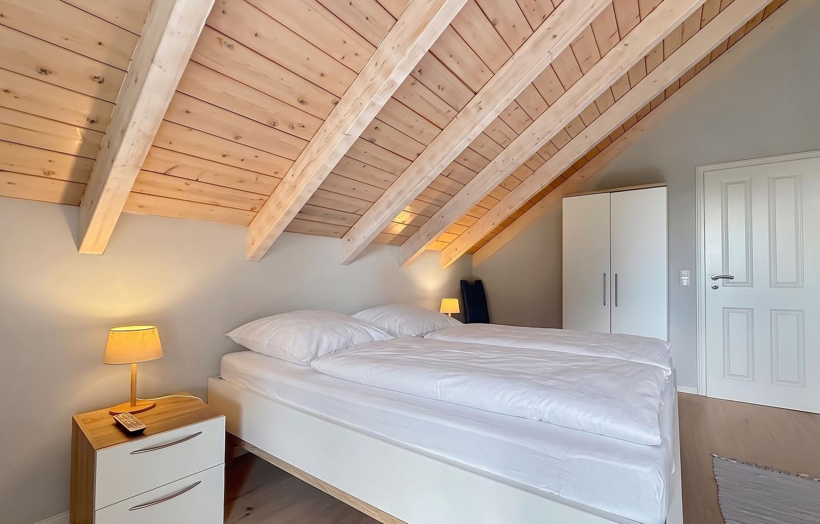 Schlafzimmer mit Doppelbett, Holzboden und schrägen Dachbalken. Neben dem Bett steht ein Nachttisch mit Lampe.
