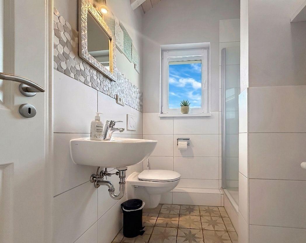 Badezimmer mit Waschbecken, Toilette und Dusche. Fenster mit Blick auf den Himmel.