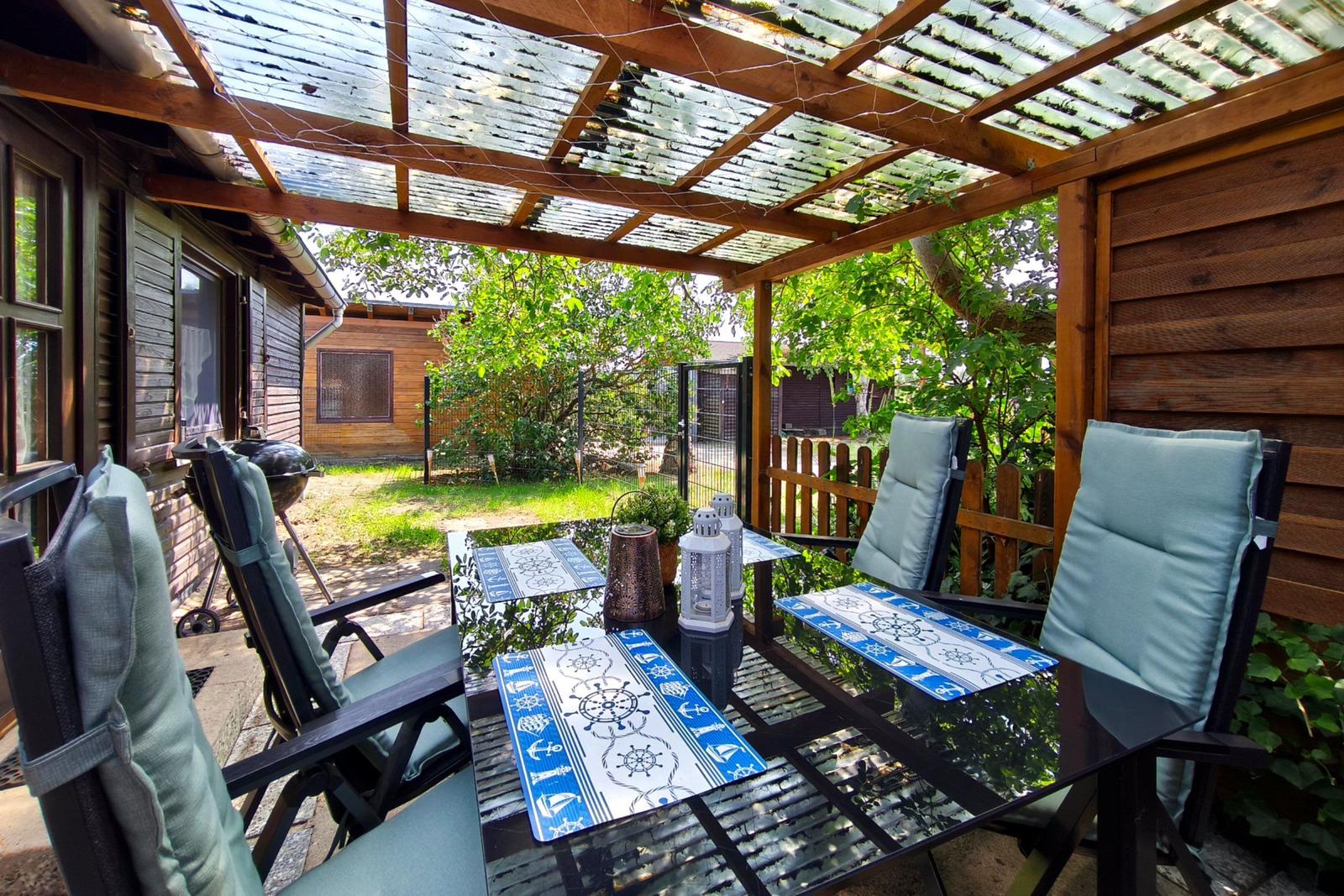 Terrasse mit Tisch, Stühlen und Grill unter Holzüberdachung mit Glasplatten.