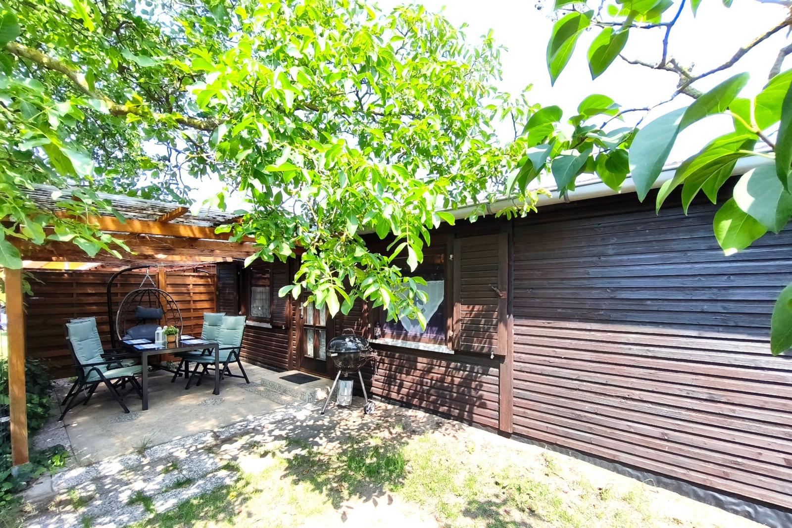 Terrasse mit Sitzgruppe, Grill und Schatten unter Bäumen vor einem Holzhaus.