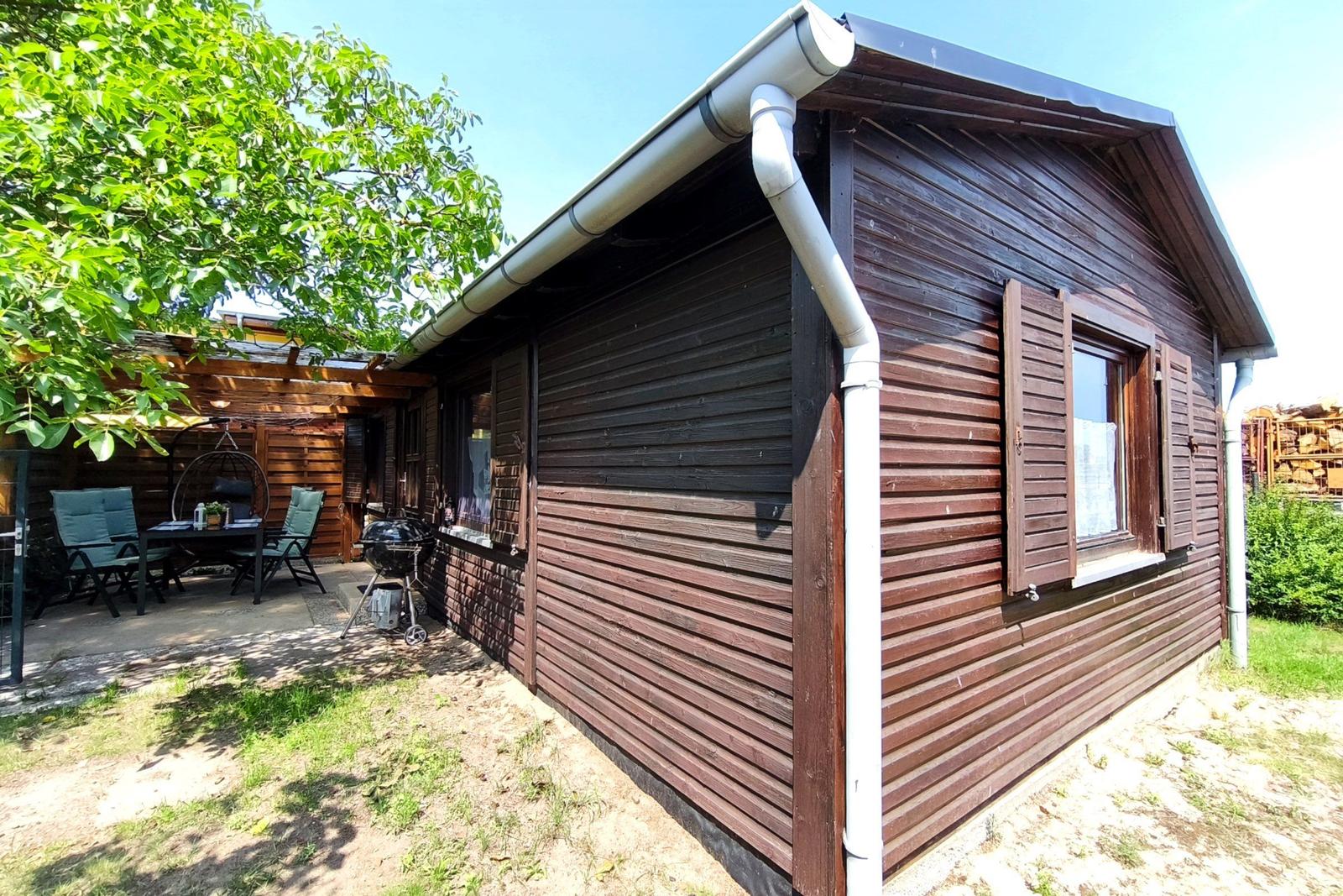 Holzhütte mit Terrasse, Grill und Sitzgelegenheiten unter Bäumen.