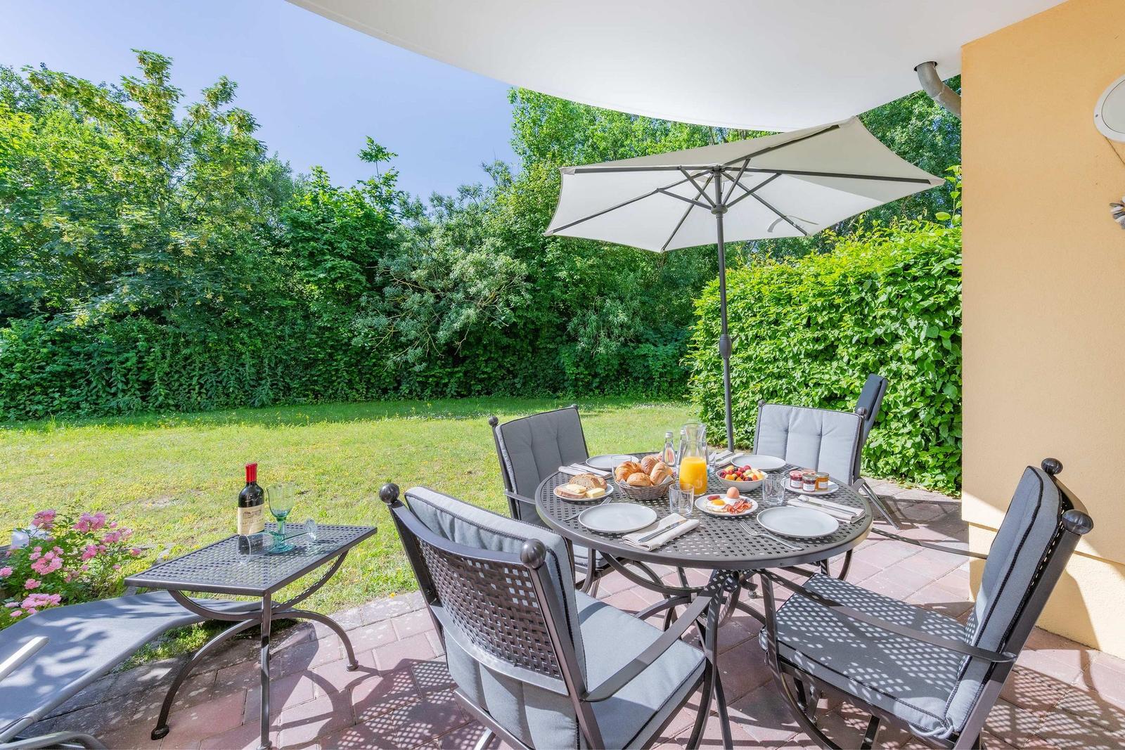 Terrasse mit Tisch, Stühlen und Sonnenschirm. Hintergrund: grüner Garten und Hecke.