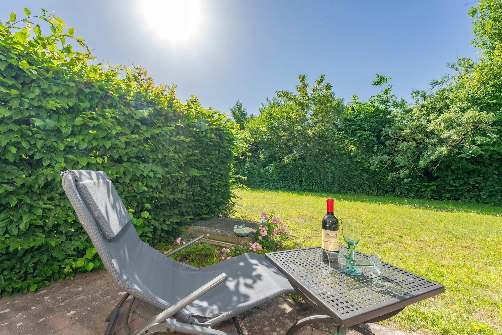 Außenterrasse mit Liegestuhl und Tisch mit Weinflasche und Gläsern. Grünhedge und Rasen im Hintergrund.