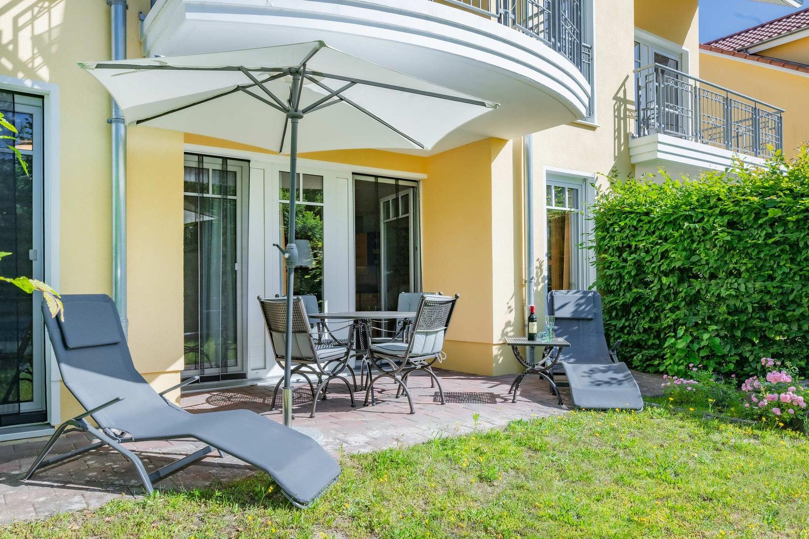 Terrasse mit Sonnenschirm, Tisch, Stühlen und Liegestühlen vor gelbem Haus