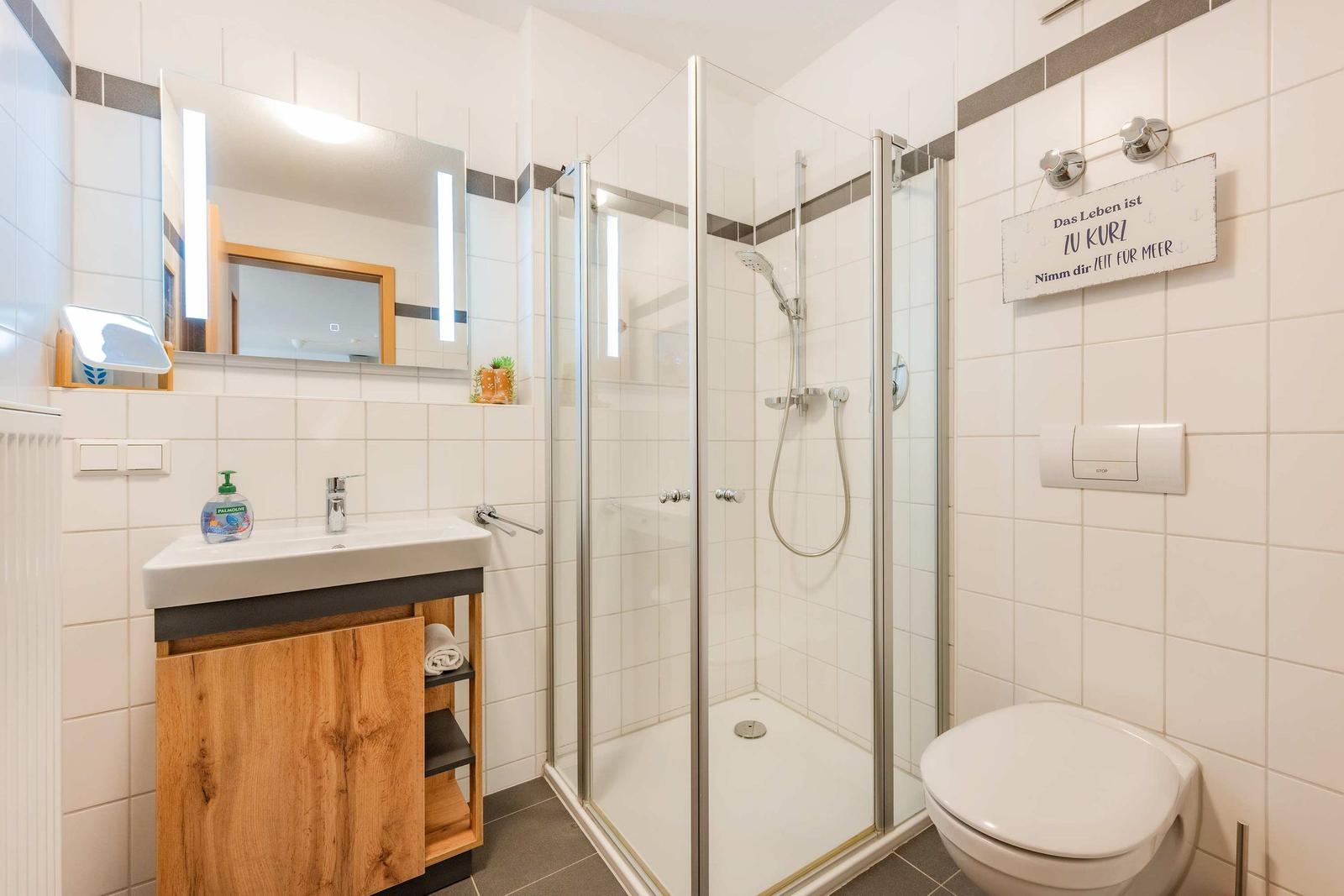 Badezimmer mit Dusche, Waschbecken und Toilette. Holzunterschrank und Spiegel mit Beleuchtung.