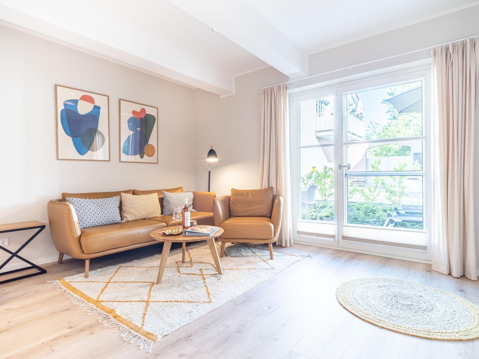 Wohnzimmer mit braunem Sofa, Sessel, Tisch und Fenster. Wandbilder und Teppiche sind sichtbar.