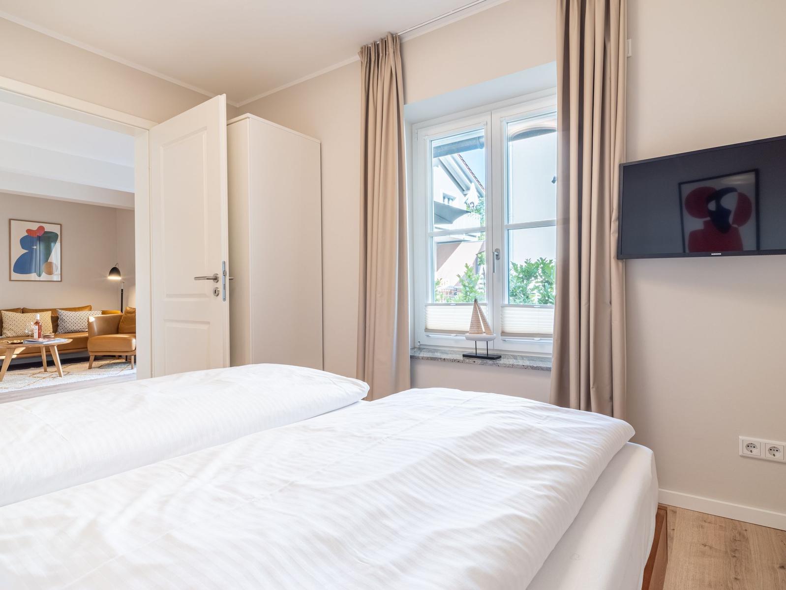Schlafzimmer mit Bett, Fenster und TV. Durchgang zur Wohnfläche mit Sofa.