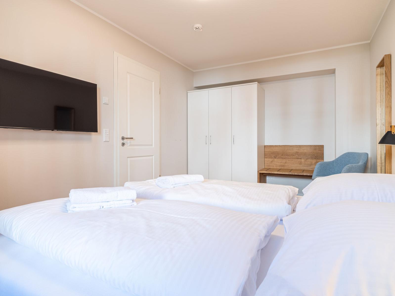 Zwei Betten mit weißen Bettwäsche, Kleiderschrank, Fernseher und Sitzgelegenheit.
