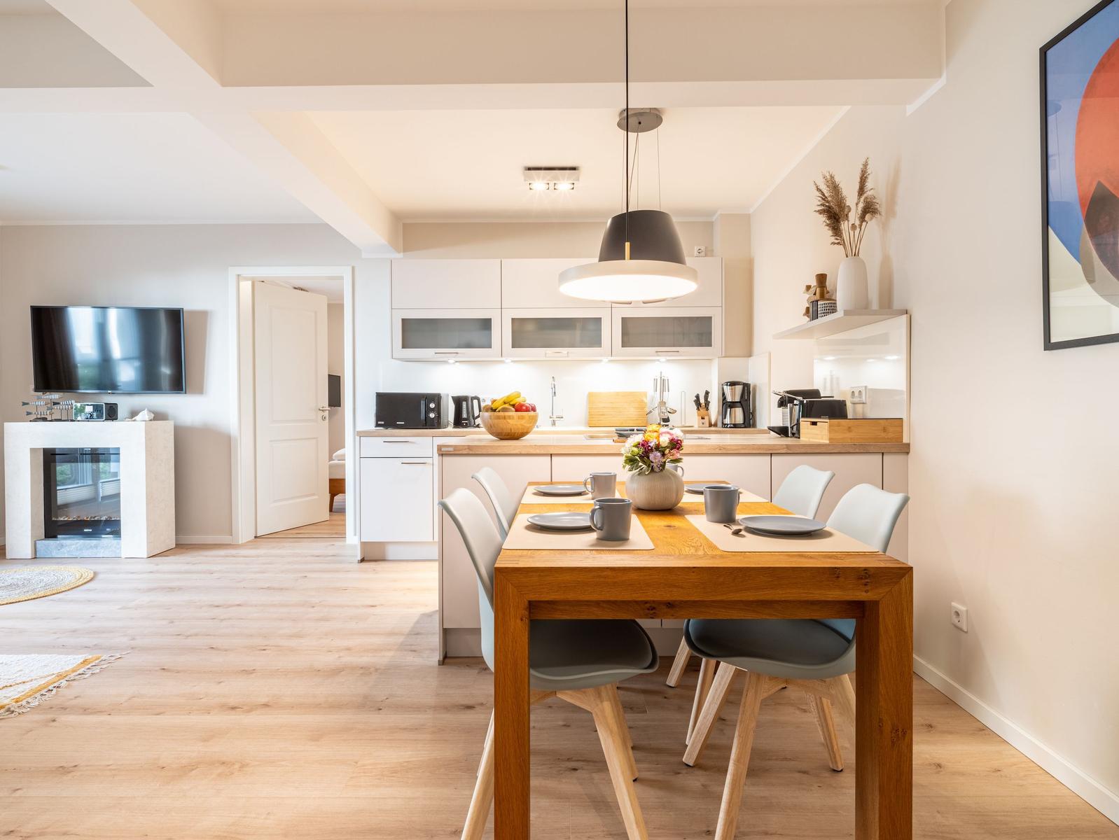 Essbereich mit Holztisch und Stühlen, Küche mit weißen Schränken und Fernseher im Wohnbereich.