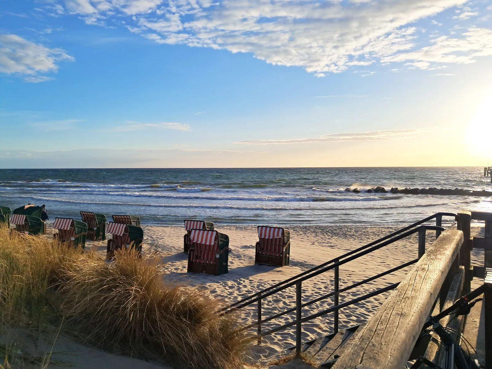 Strand mit Liegestühlen und Holzsteg. Sonne geht unter. Wellen brechen am Horizont.