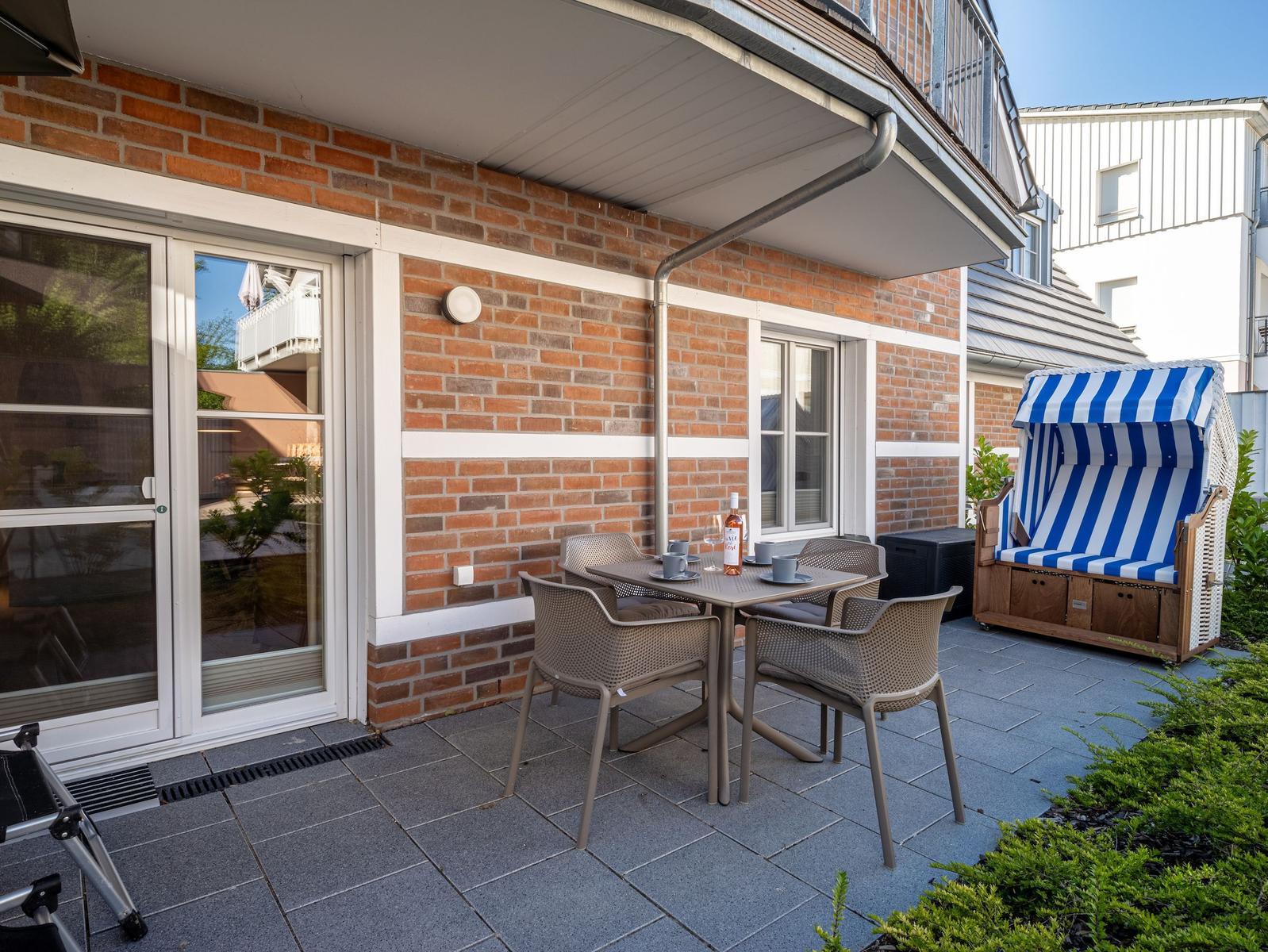 Terrasse mit Tisch, Stühlen und Strandkorb. Hintergrund: Backsteinmauer und Glas-Tür.