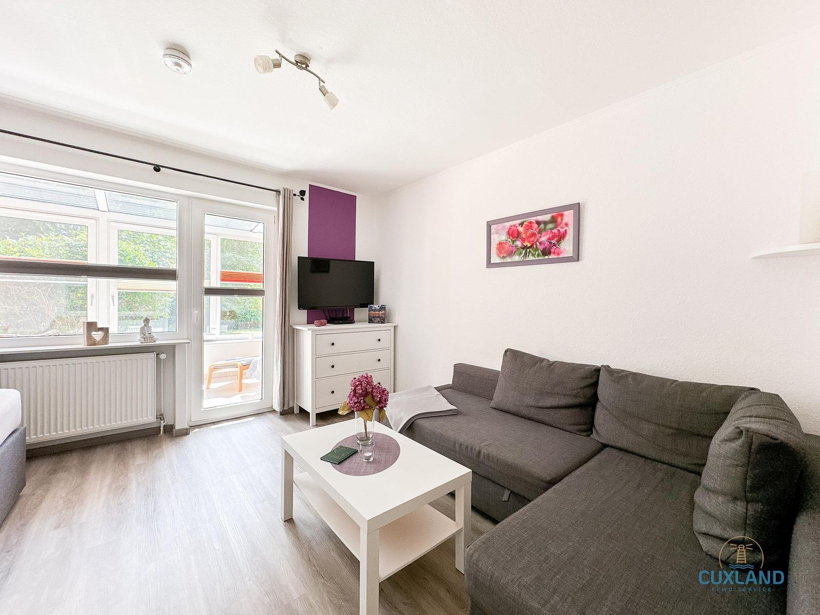 Wohnzimmer mit grauem Sofa, weißem Tisch und TV auf Schrank. Fenster und Tür zur Terrasse.