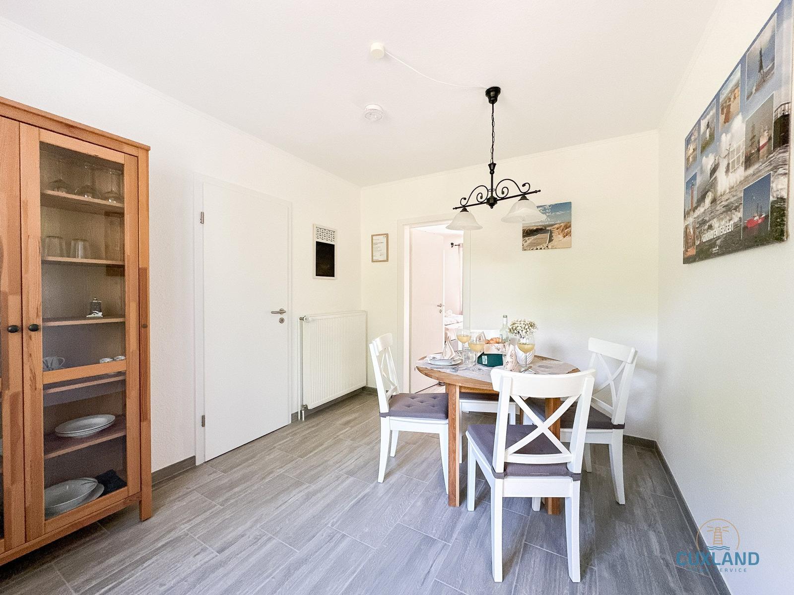 Esszimmer mit Holztisch, weißen Stühlen und Glasvitrine. Wandbild zeigt Küstenlandschaft.