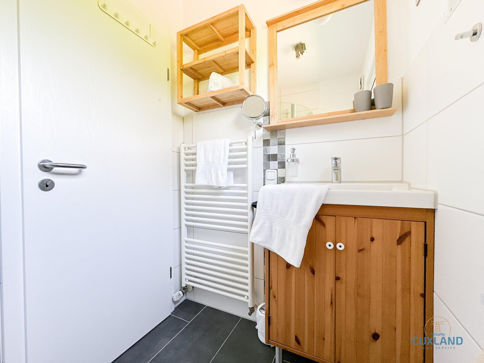 Badezimmer mit Holzschrank, Spiegel und Heizkörper. Weiße Wände und dunkler Boden.