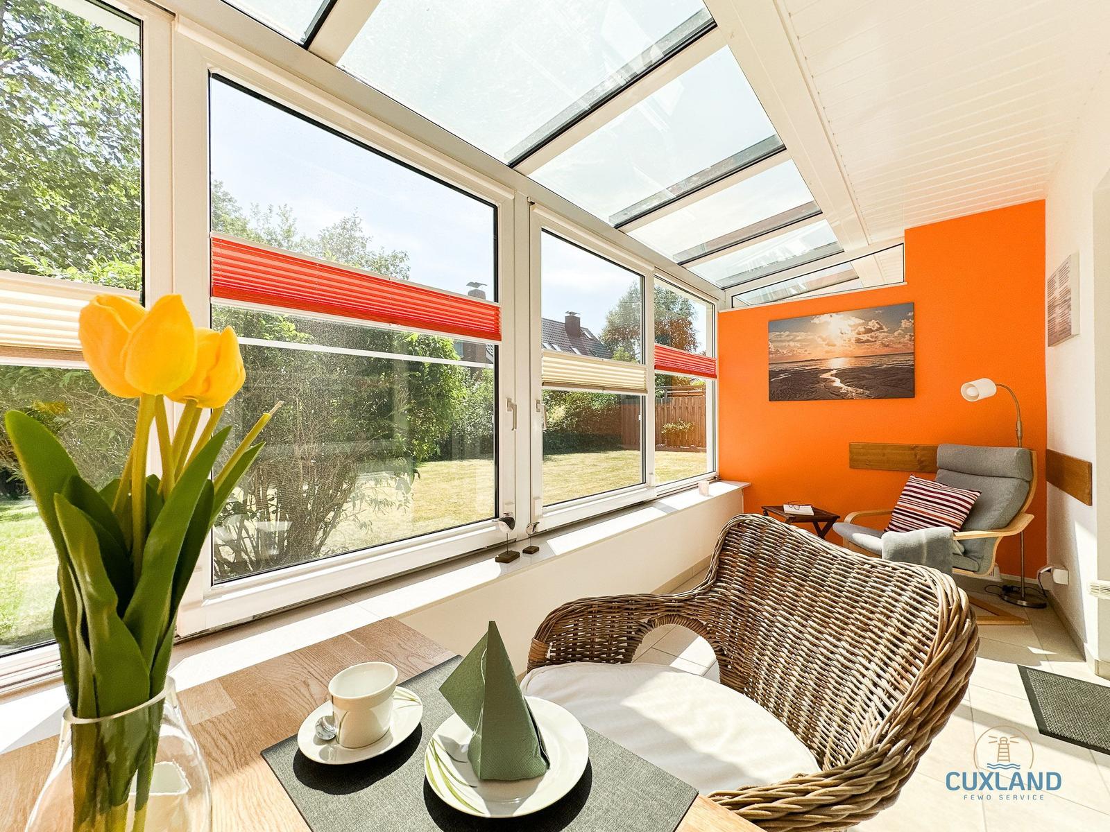 Terrasse mit Sitzgelegenheiten und Blick auf den Garten. Orange Wand und Fenster mit roten Markisen.