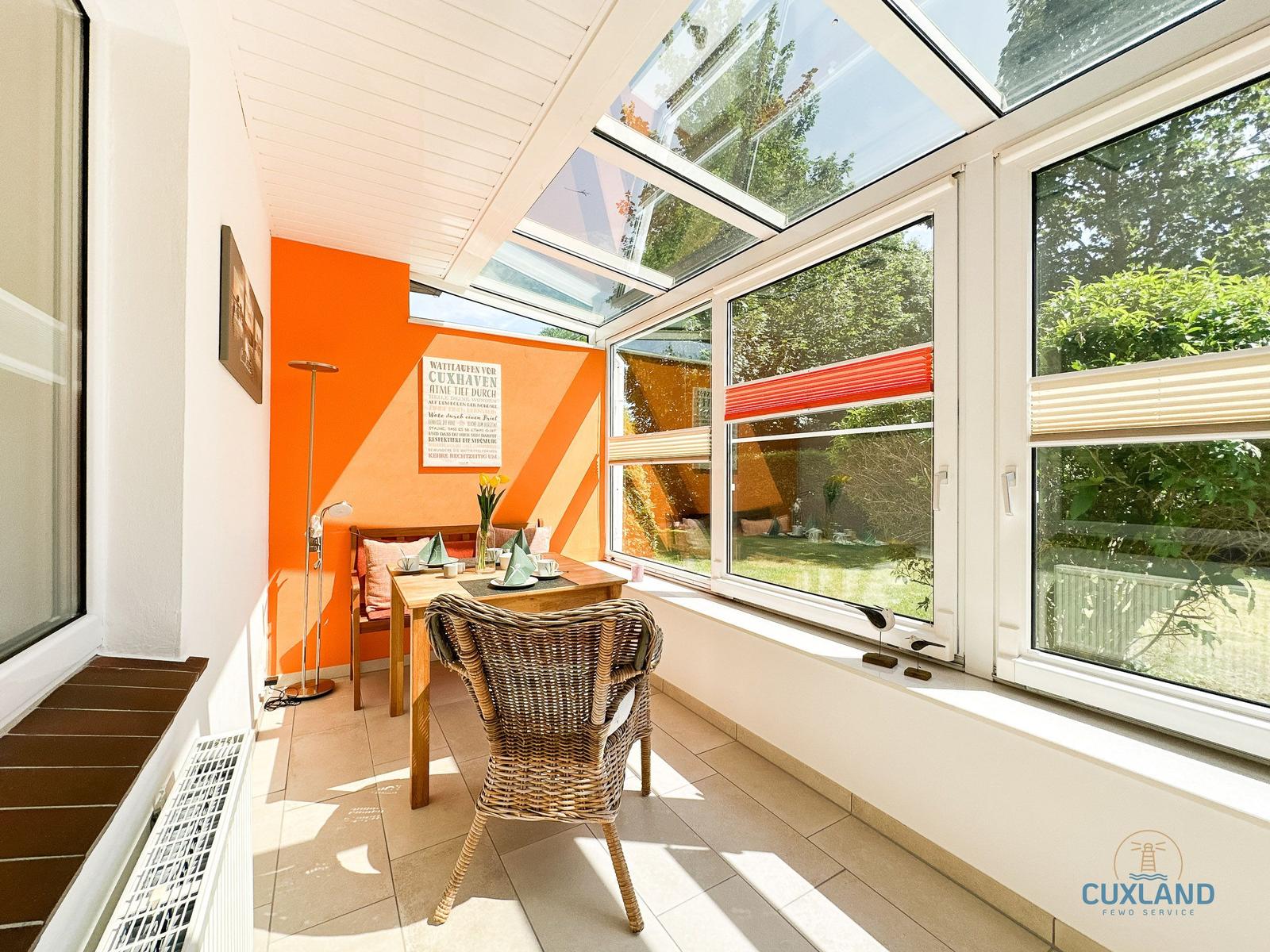 Sonniger Raum mit Sitzgelegenheit, Tisch und orangefarbener Wand. Große Fenster und Dachfenster mit Blick auf Garten.