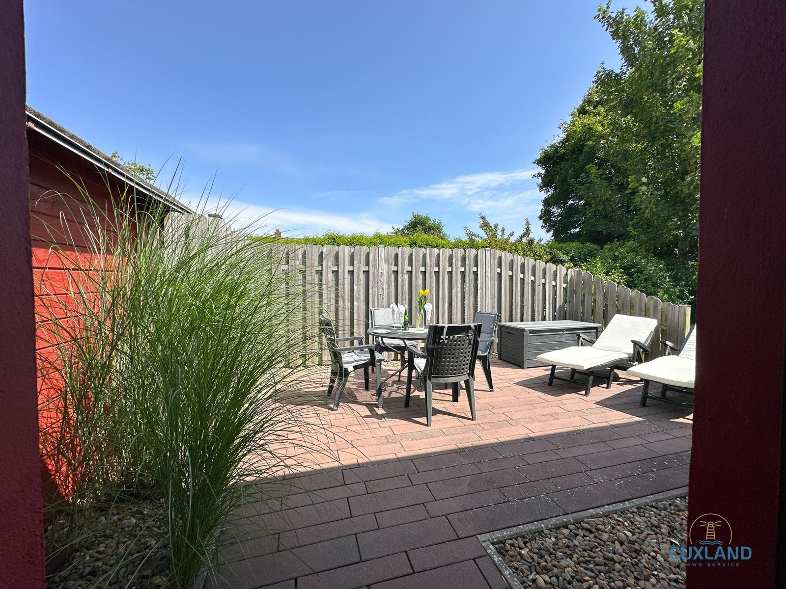 Terrasse mit Tisch, Stühlen und Liegen. Holzzaun und grüne Pflanzen umgeben den Außenbereich.