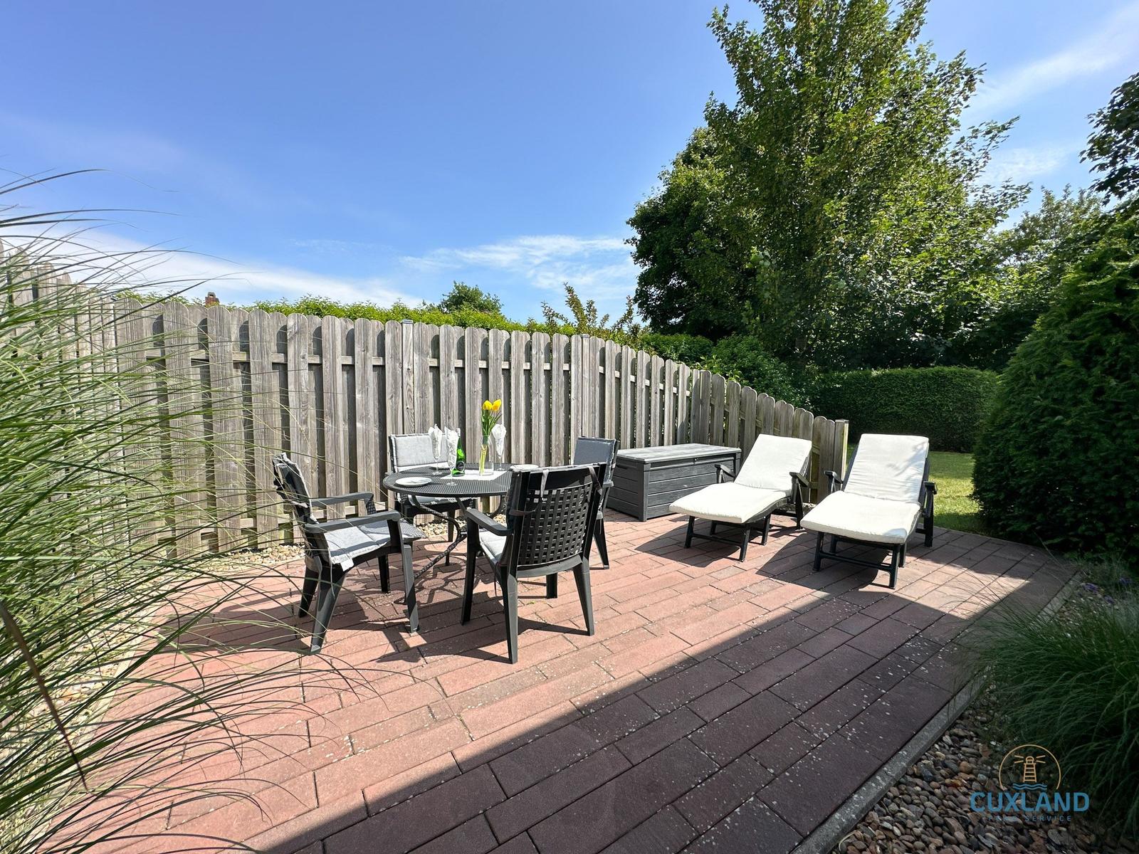 Terrasse mit Tisch, Stühlen und Liegestühlen. Hinterhof mit Holzzaun und Bäumen.
