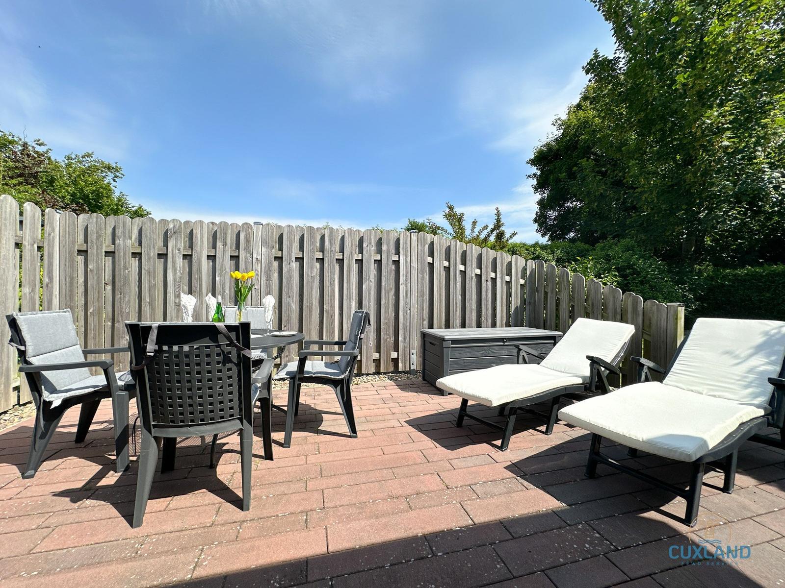 Terrasse mit Tisch, Stühlen und Liegen. Hintergrund: Holzzaun und Bäume.